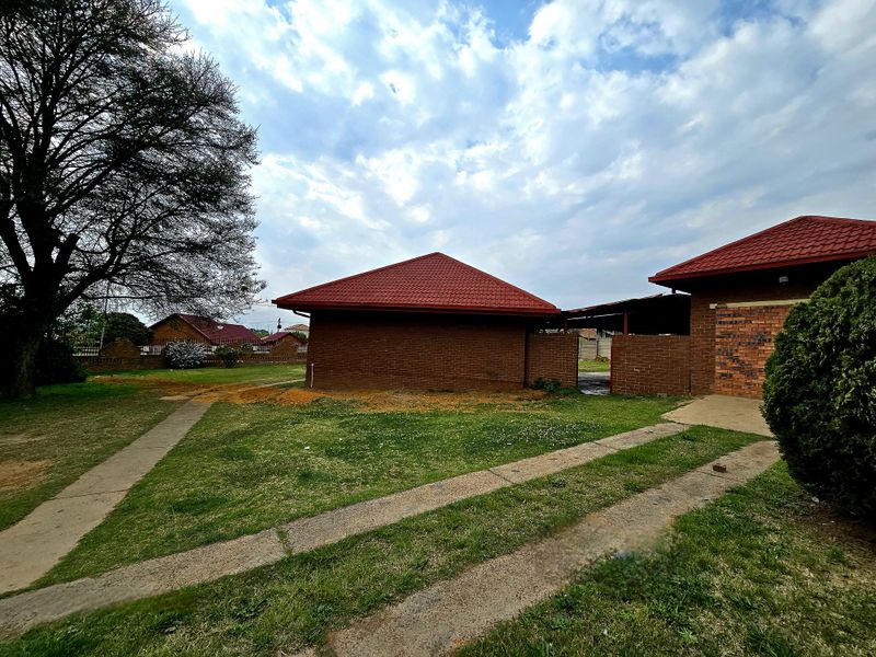 Rear view of the property featuring a brick exterior, red tiled roof, and a spacious garden area with concrete pathways and driveway.
