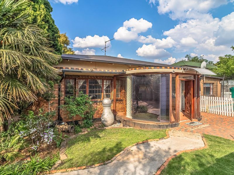Rear view of the property featuring a brick facade, a unique curved glass sunroom, a paved patio area, and a well-maintained garden with a picket fence.