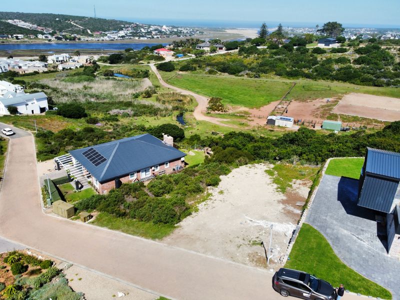 Aerial view of a vacant residential plot situated in a coastal neighborhood, featuring paved street access and proximity to established homes and natural landscape.