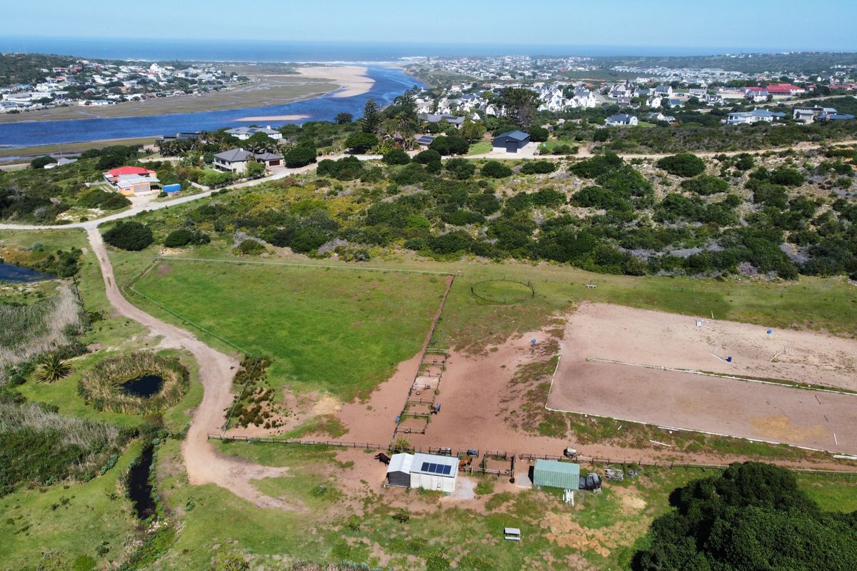 Aerial view of a residential vacant land property featuring equestrian facilities, fenced paddocks, and outbuildings with solar panels, overlooking a coastal landscape.
