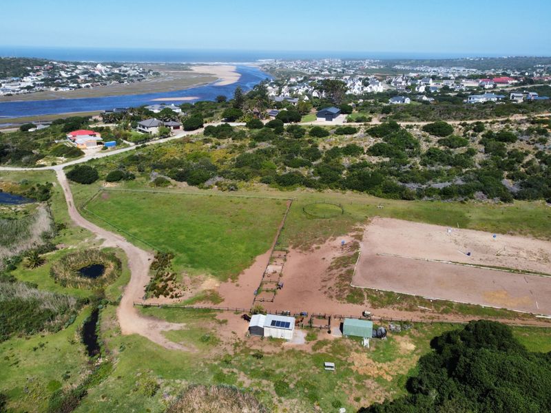 Aerial view of a residential vacant land property featuring equestrian facilities, fenced paddocks, and outbuildings with solar panels, overlooking a coastal landscape.