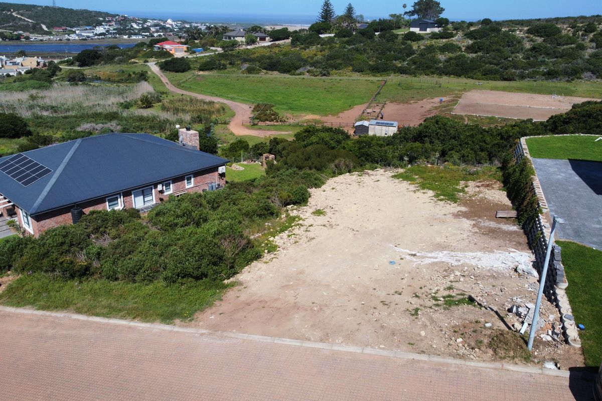 Aerial view of a vacant residential plot featuring a paved road frontage and adjacent retaining wall, situated in a coastal landscape.