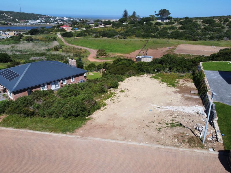 Aerial view of a vacant residential plot featuring a paved road frontage and adjacent retaining wall, situated in a coastal landscape.