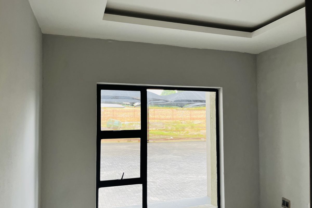 A bright, empty bedroom featuring a modern recessed ceiling design and a large black aluminium window frame overlooking the exterior area.