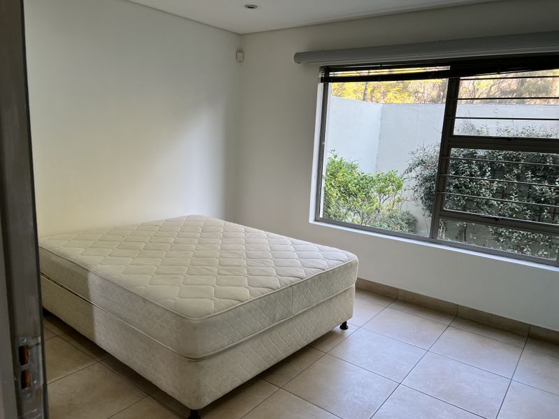 A bright, neutral bedroom featuring tiled flooring, recessed lighting, and a large window with security bars overlooking a private garden area.