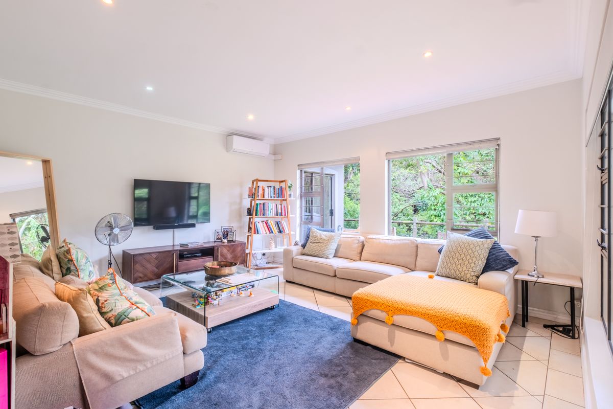 A bright and spacious living room featuring tiled flooring, recessed downlighting, and large windows with views of the surrounding greenery.
