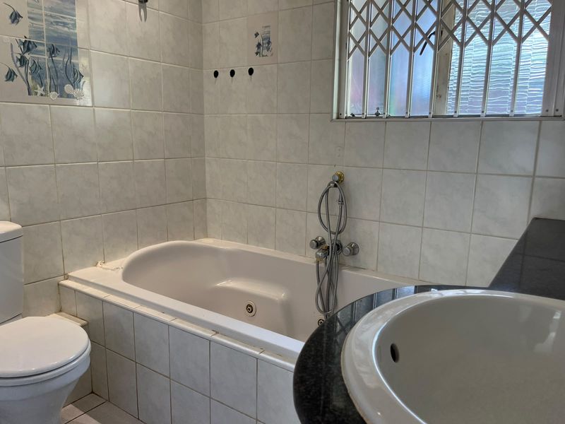 A functional bathroom featuring a built-in bathtub, vanity basin, and toilet, finished with tiled walls and a window with security bars.