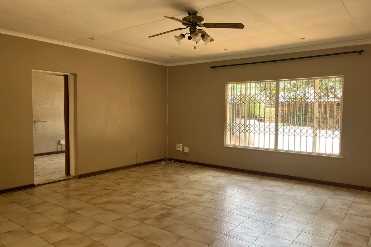 A spacious, empty living room featuring tiled flooring, a ceiling fan, and a large window with a security gate.