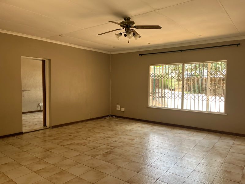 A spacious, empty living room featuring tiled flooring, a ceiling fan, and a large window with a security gate.