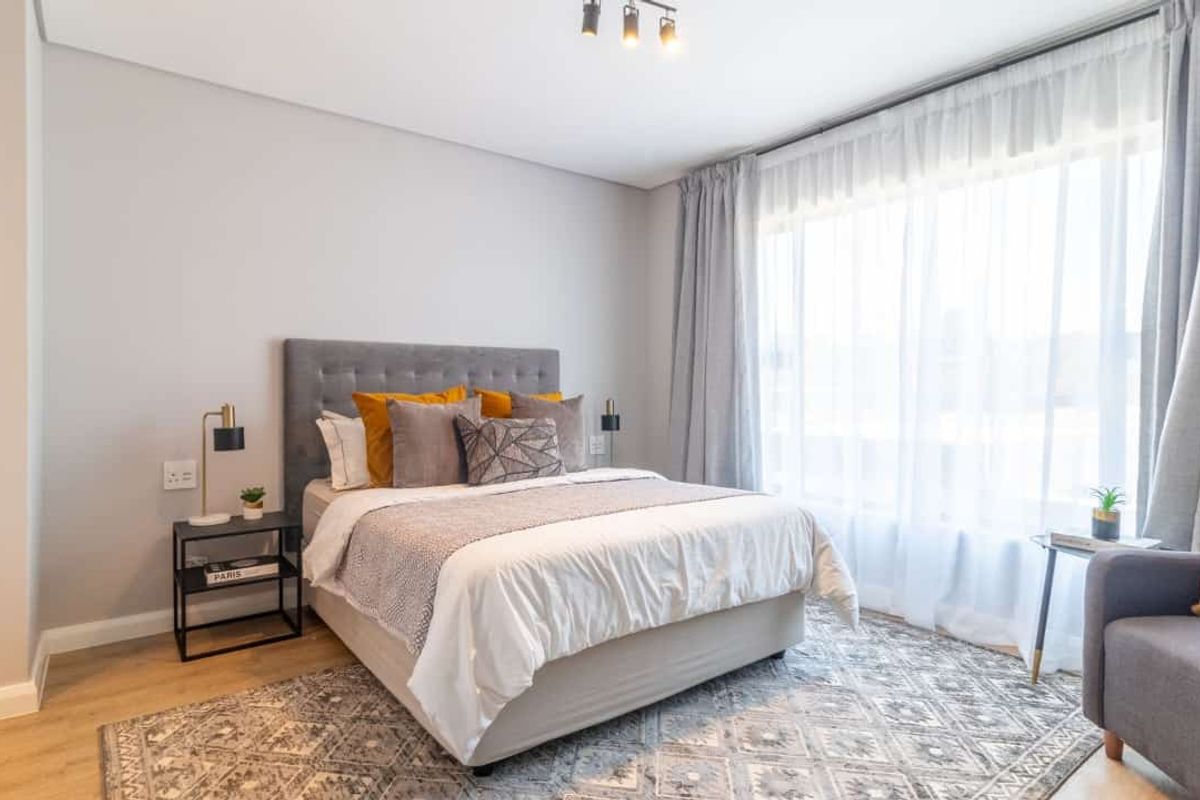 A well-lit, modern bedroom featuring neutral wall tones, laminate flooring, and a large window with sheer curtains.