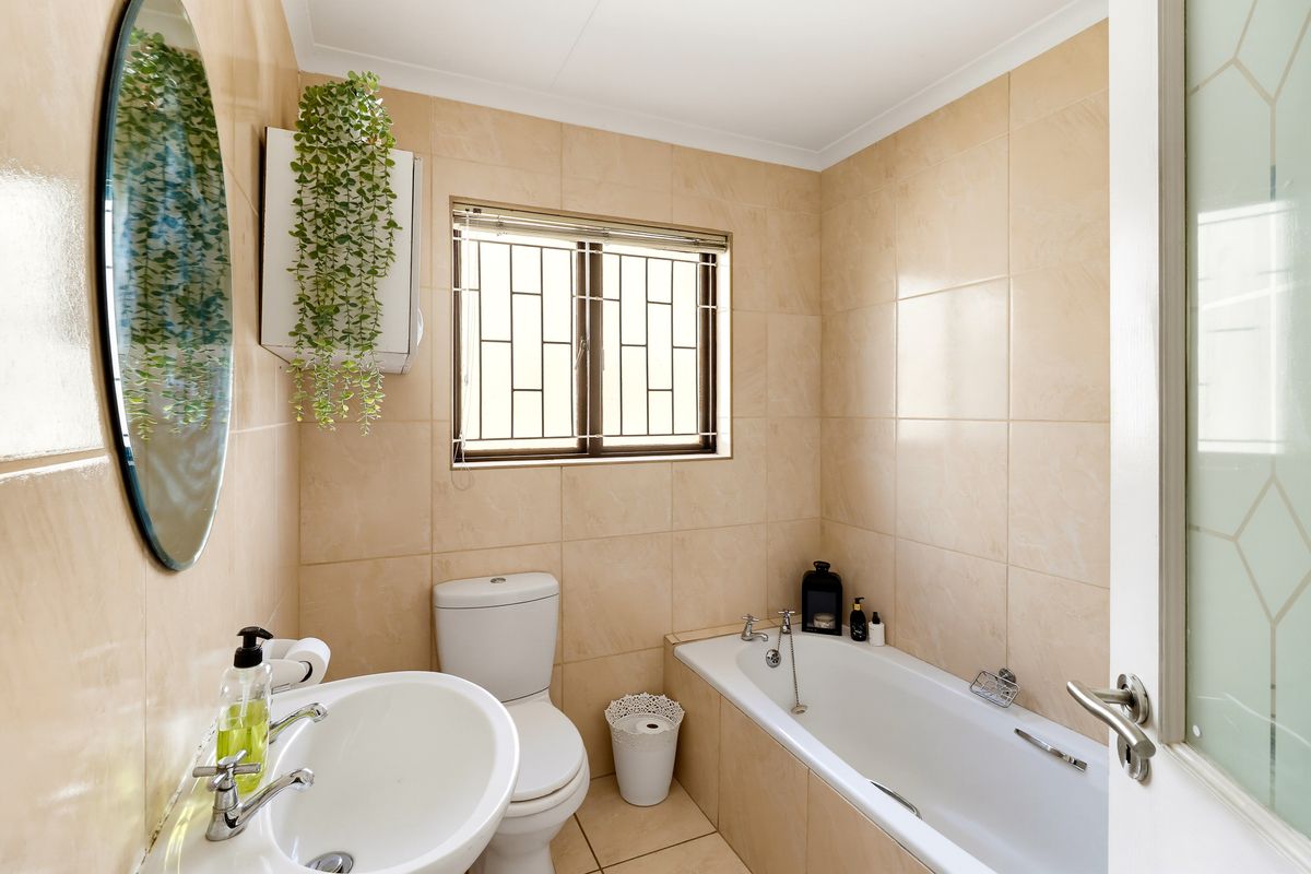 A functional bathroom featuring tiled walls and flooring, a built-in bathtub, a pedestal basin, and a window with security bars.