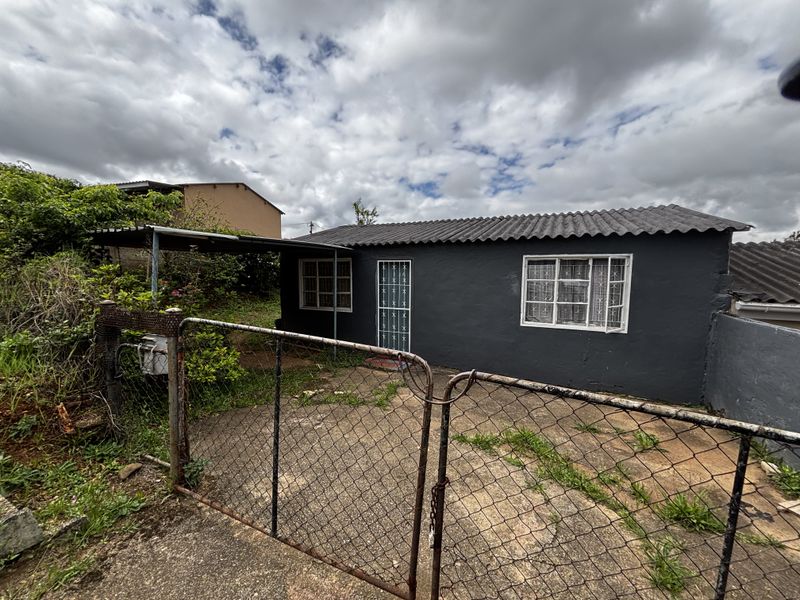 Front exterior view of a townhouse featuring a dark painted facade, corrugated roof, security gate, and a concrete driveway with a carport.