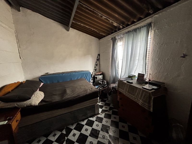 A bedroom featuring exposed corrugated roofing, tiled flooring, and a window with security bars, requiring significant cosmetic updates and repairs to wall finishes.