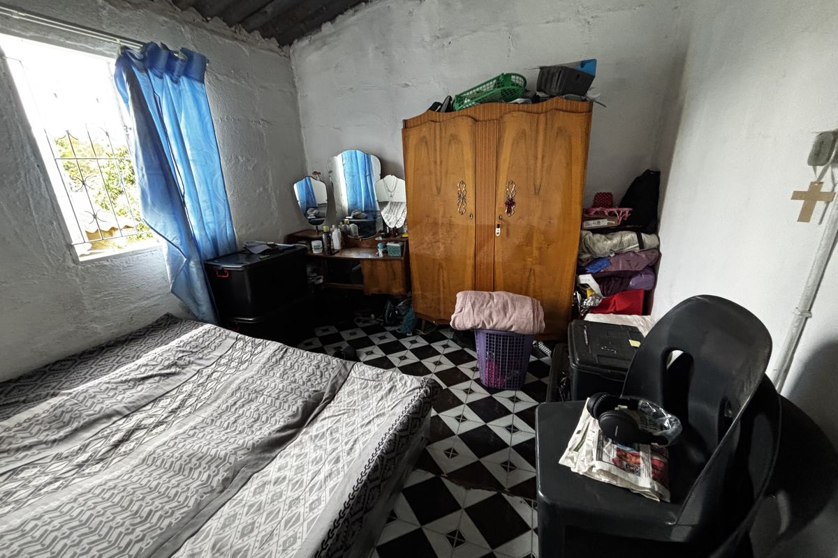 A bedroom featuring exposed roof rafters, checkered floor tiles, and a window with security bars, requiring cosmetic updates to improve the overall finish.