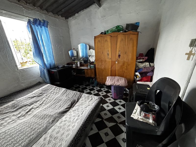 A bedroom featuring exposed roof rafters, checkered floor tiles, and a window with security bars, requiring cosmetic updates to improve the overall finish.