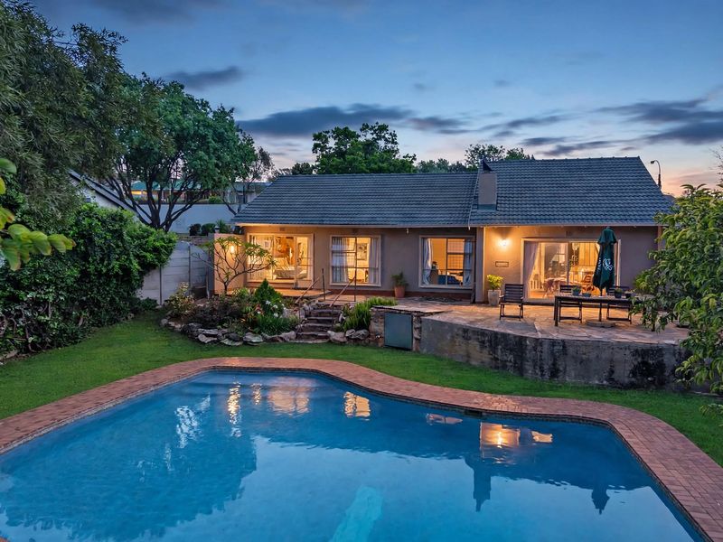 Rear view of the property featuring a swimming pool with brick coping, a stone patio area, and a well-maintained garden at dusk.