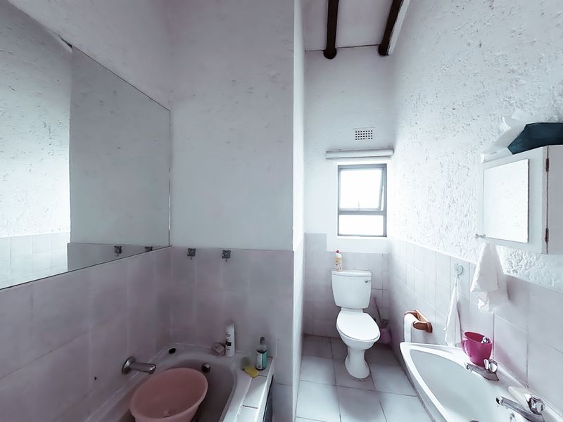 A functional bathroom featuring a built-in bathtub, pedestal basin, toilet, and large wall-mounted mirror, finished with tiled surfaces and exposed ceiling beams.
