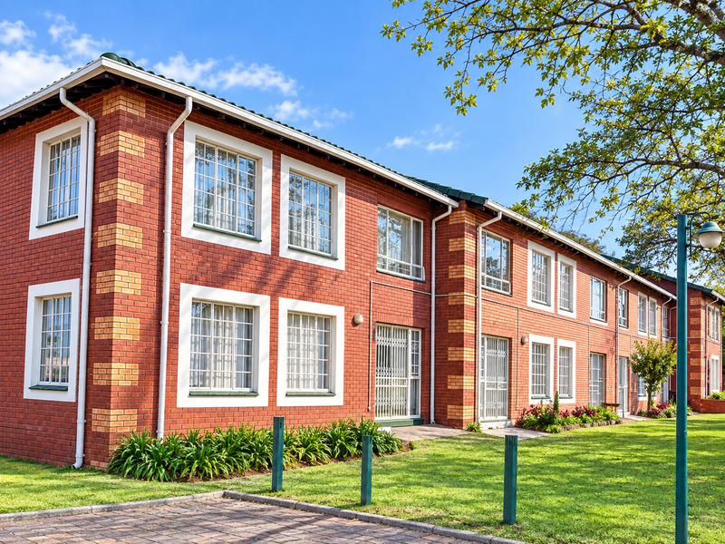 A well-maintained apartment building exterior featuring classic red brickwork, white window frames, security gates, and a neatly landscaped garden area.