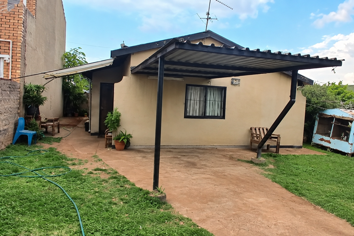 Rear view of the property featuring a metal carport, concrete driveway, and a brick boundary wall in a garden setting.