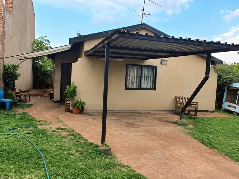 Rear view of the property featuring a metal carport, concrete driveway, and a brick boundary wall in a garden setting.