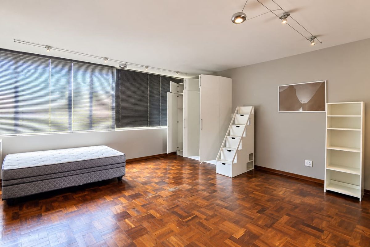 Spacious bedroom featuring classic parquet flooring, built-in storage cupboards, and large windows with venetian blinds, offering a bright and functional living space.