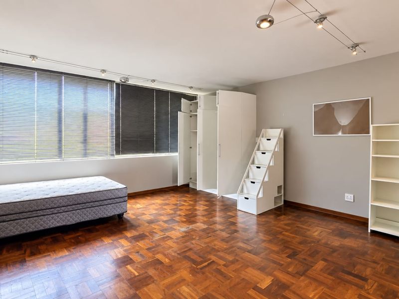 Spacious bedroom featuring classic parquet flooring, built-in storage cupboards, and large windows with venetian blinds, offering a bright and functional living space.