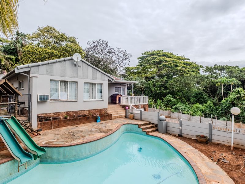Rear exterior view featuring a swimming pool with stone paving, brick retaining walls, and a surrounding fence set against a lush garden backdrop.