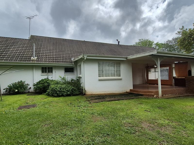 Rear view of the house featuring a covered patio with a built-in brick braai, overlooking a spacious garden with a tiled roof structure.