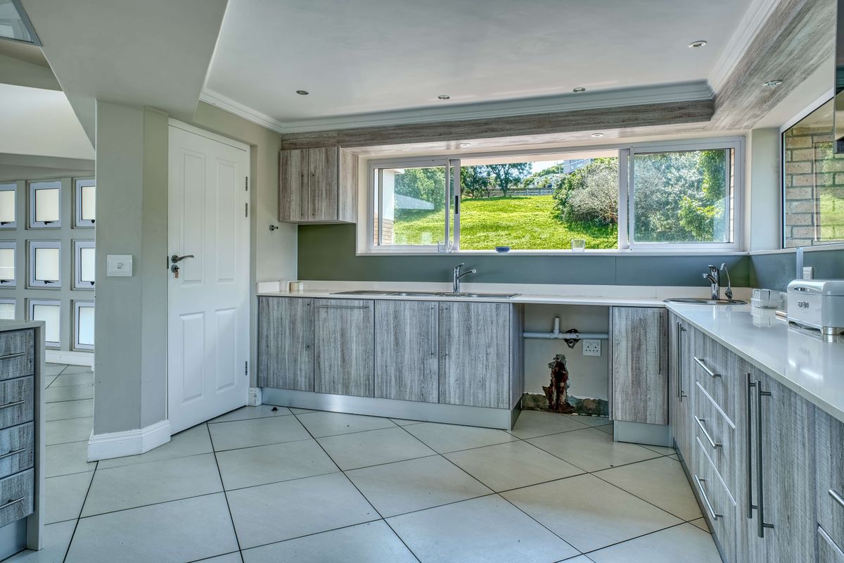 A kitchen featuring wood-look cabinetry, tiled floors, and a large window with garden views. Requires repair to the wall and plumbing area under the sink.