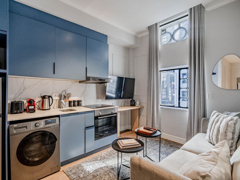 Modern open-plan apartment featuring a compact, well-equipped kitchen with blue cabinetry, integrated appliances, and a bright living area with tall windows.