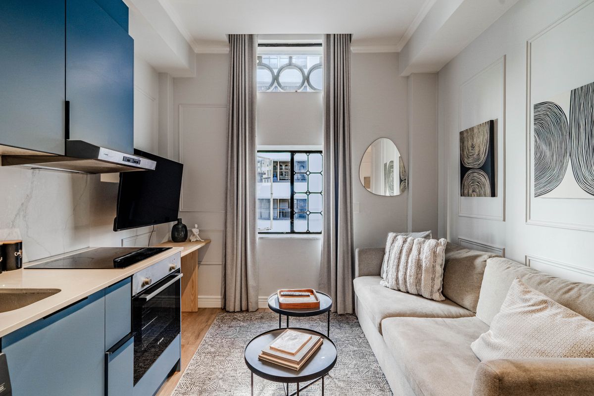 A modern open-plan apartment living area featuring a compact, well-equipped kitchen with blue cabinetry and a bright, stylish lounge space.