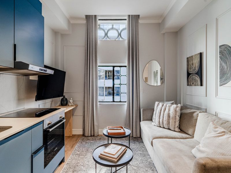 A modern open-plan apartment living area featuring a compact, well-equipped kitchen with blue cabinetry and a bright, stylish lounge space.