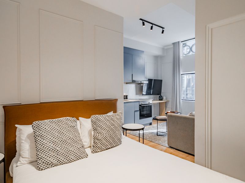 Modern open-plan apartment featuring a bedroom area with decorative wall paneling, flowing into a compact kitchen with sleek blue cabinetry and integrated appliances.