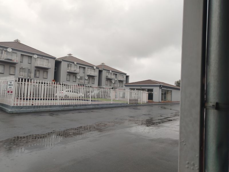 Exterior view of a multi-story apartment complex featuring paved driveway access, security fencing, and residential balconies under a cloudy sky.