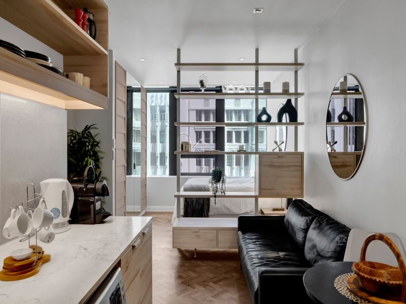 Modern open-plan studio apartment featuring herringbone timber flooring, a sleek kitchen with stone countertops, and a functional built-in room divider.