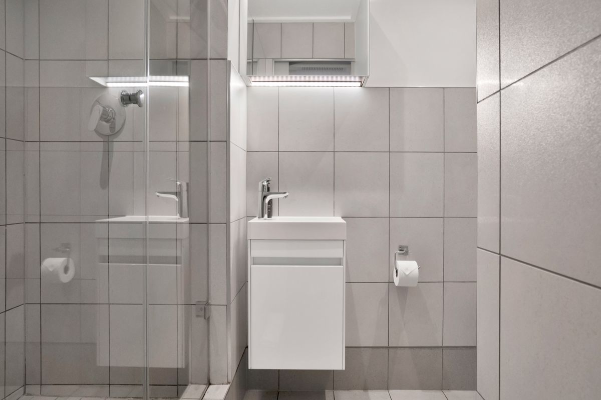 Modern, compact bathroom featuring neutral grey tiling, a wall-mounted vanity with basin, and a glass-enclosed shower area with integrated lighting.