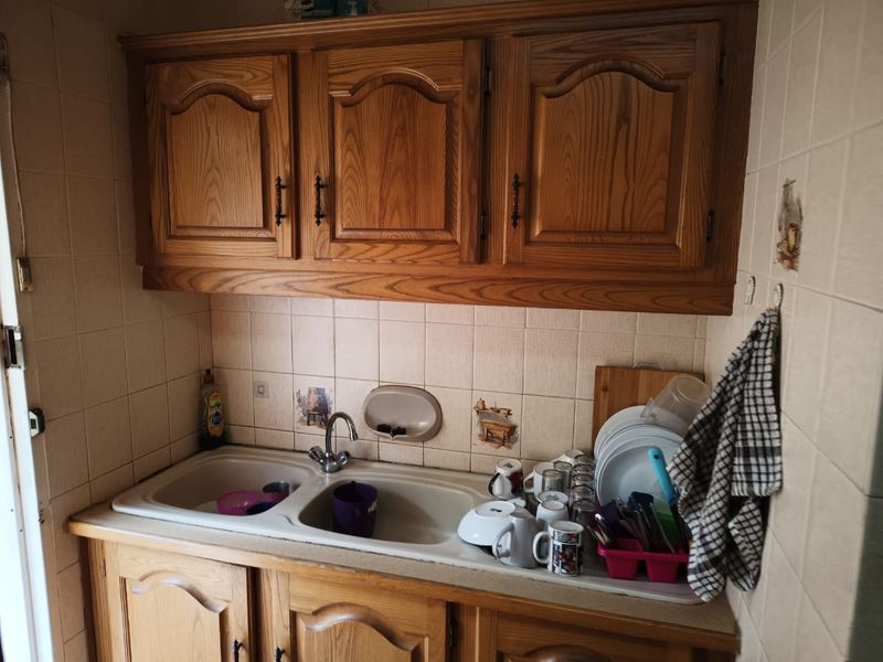 A compact kitchen area featuring traditional wooden cabinetry, a double basin sink, and tiled backsplash, offering a functional space with potential for modernization.