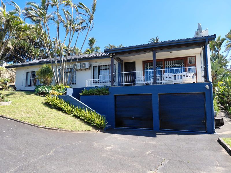 Front elevation of a residential house featuring a double garage, elevated covered patio, and landscaped garden area with mature palm trees.