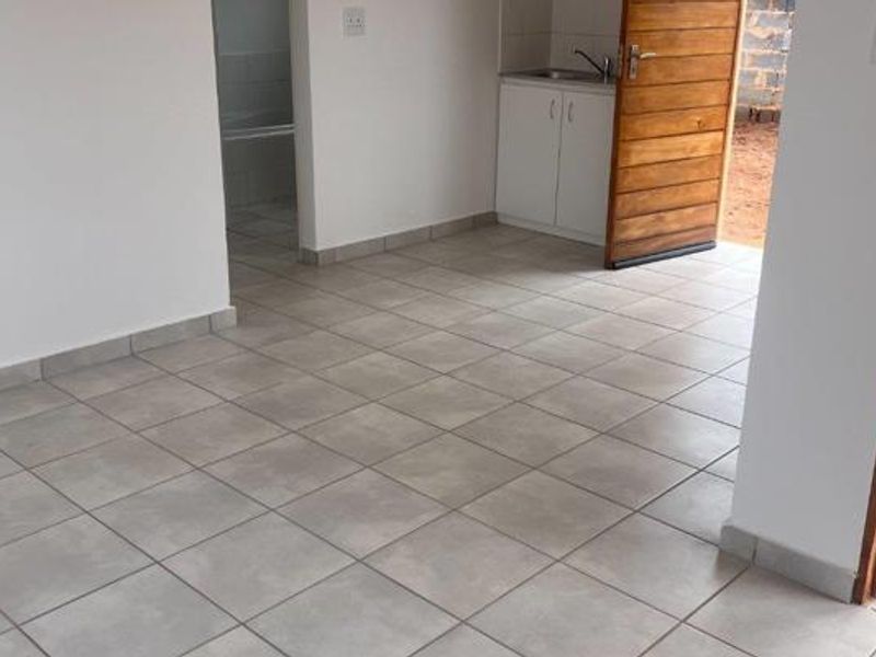 An open-plan living area featuring neutral tiled flooring, a compact kitchenette with built-in cabinetry, and direct access to an outdoor space.