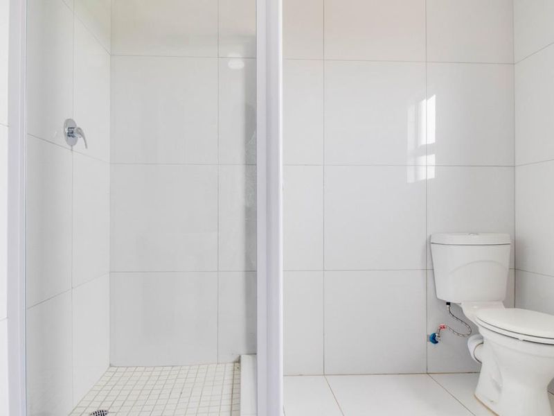 A clean, minimalist bathroom featuring white wall and floor tiles, a walk-in shower enclosure, and a standard toilet.