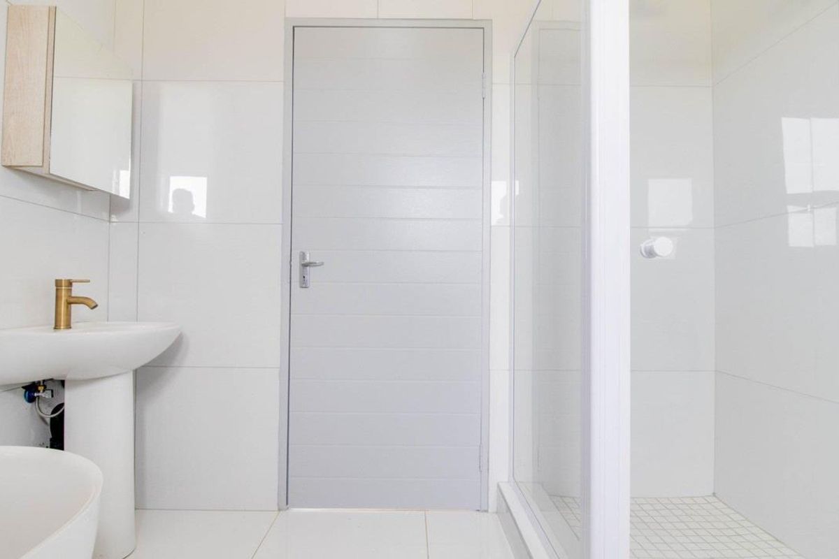 A clean, modern bathroom featuring white wall tiles, a pedestal basin with brass tapware, and a glass-enclosed shower.