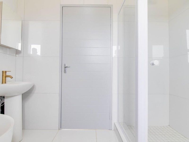A clean, modern bathroom featuring white wall tiles, a pedestal basin with brass tapware, and a glass-enclosed shower.