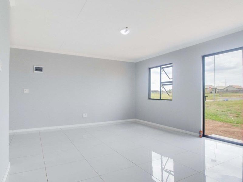 A bright, empty living room featuring modern white tiled flooring, neutral grey walls, and a large sliding glass door leading to the exterior.