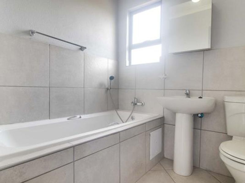 A clean, functional bathroom featuring a bathtub with handheld shower mixer, pedestal basin, toilet, and wall-mounted mirror cabinet with neutral wall and floor tiling.