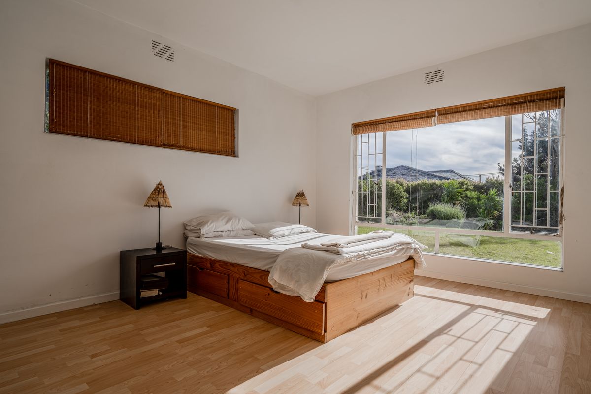 A bright bedroom featuring light laminate flooring, large windows with security bars, and natural light, offering a peaceful view of the garden.