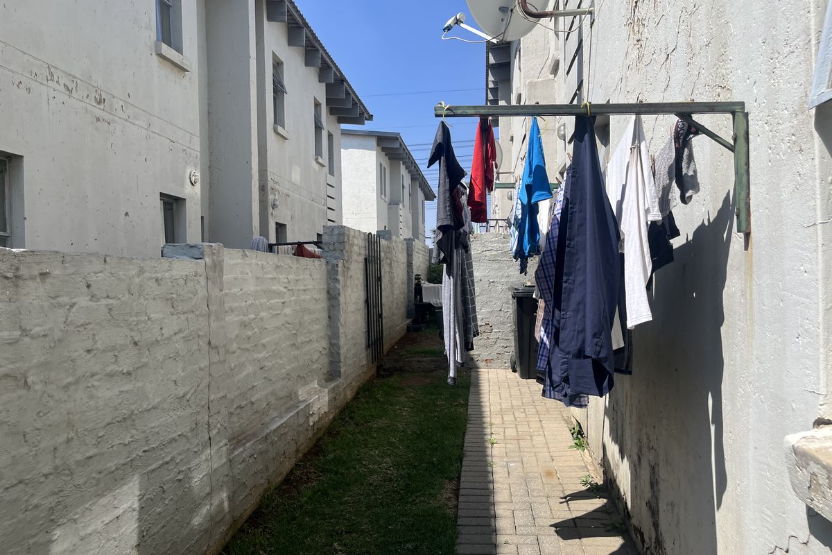 A narrow rear exterior side passage featuring a paved walkway, brick boundary wall, and wall-mounted clothesline, showing signs of weathered exterior finishes.