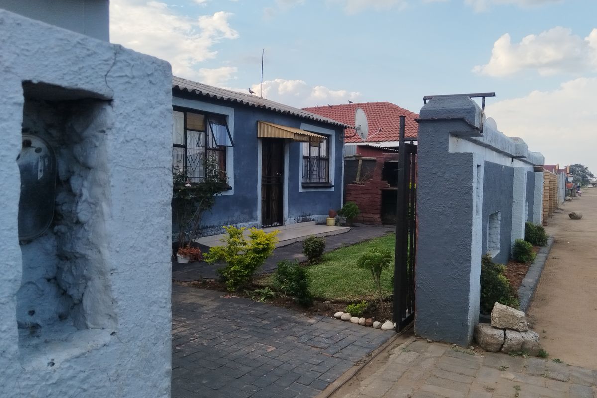 Front view of a residential property featuring a paved driveway, boundary wall with gate, and a small front garden with a built-in brick braai.