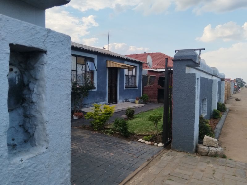 Front view of a residential property featuring a paved driveway, boundary wall with gate, and a small front garden with a built-in brick braai.