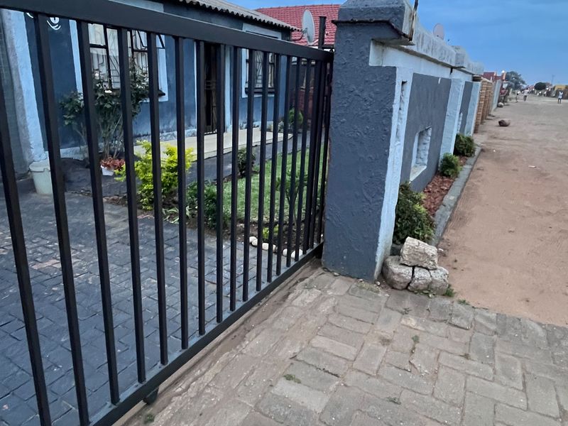 Front view of the property featuring a secure metal sliding gate, perimeter wall, and paved driveway leading to the house entrance.