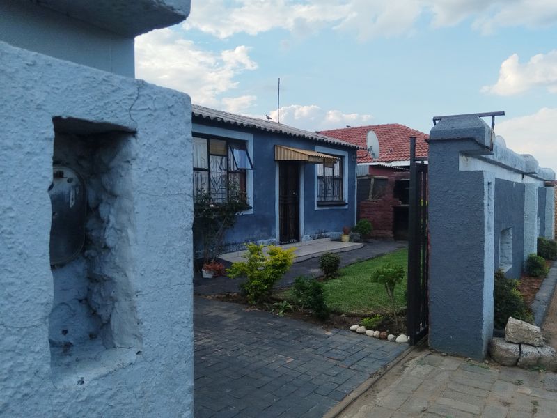 Front view of a residential property featuring a paved driveway, security gate, boundary wall, and a house with a tiled roof and security bars.